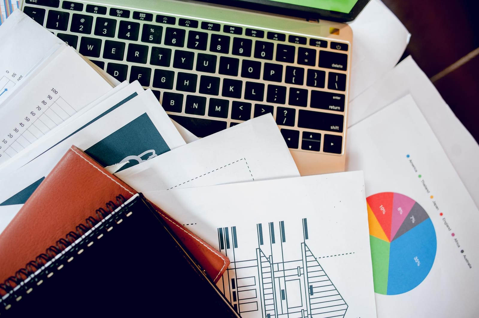Blick von oben auf eine Laptoptastatur, bedeckt mit gedruckten Diagrammen, einem Kreisdiagramm und Notizbüchern.